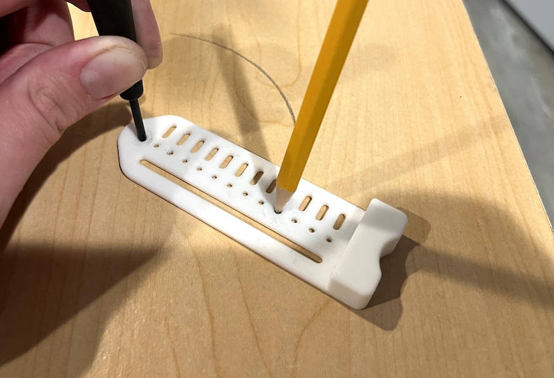 May include: A white plastic marking guide with a series of holes and slots is used with a pencil and a black marking tool on a wooden surface. The pencil is yellow with a gray tip.