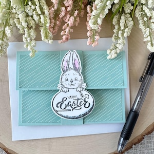 May include: A light blue card with a white bunny holding a sign that says "Happy Easter". The card has a striped pattern.