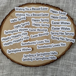 May include: A collection of white stickers with black text, featuring Easter and Spring greetings. Phrases include "Wishing You a Blessed Easter," "Happy Easter," and "Hello Spring." The stickers are arranged on a rustic wooden slice.