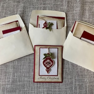 Pode incluir: Tr&ecirc;s envelopes brancos com cart&otilde;es vermelhos e marrons dentro. O cart&atilde;o na frente tem um ornamento vermelho com um la&ccedil;o verde e o texto "Merry Christmas".