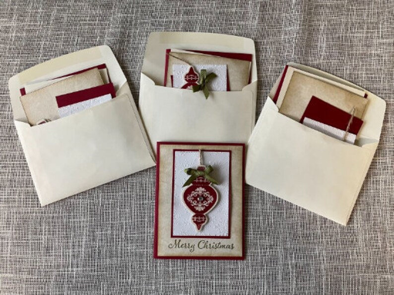Pode incluir: Tr&ecirc;s envelopes brancos com cart&otilde;es vermelhos e marrons dentro. O cart&atilde;o na frente tem um ornamento vermelho com um la&ccedil;o verde e o texto "Merry Christmas".