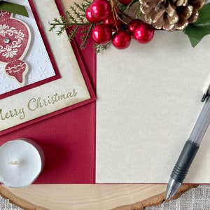 Pode incluir: Um cart&atilde;o de Natal vermelho e branco com um ornamento dourado e o texto "Merry Christmas". O cart&atilde;o est&aacute; sobre uma superf&iacute;cie de madeira com uma vela branca, uma caneta e um ramo de azevinho com bagas vermelhas.