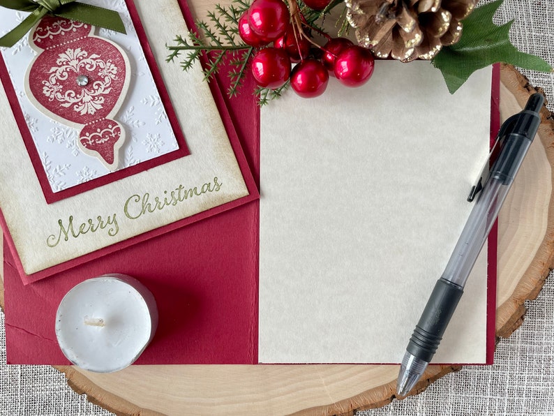 Pode incluir: Um cart&atilde;o de Natal vermelho e branco com um ornamento dourado e o texto "Merry Christmas". O cart&atilde;o est&aacute; sobre uma superf&iacute;cie de madeira com uma vela branca, uma caneta e um ramo de azevinho com bagas vermelhas.