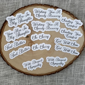 May include: A collection of white stickers with black script on a wooden slice. The stickers feature phrases like "You Are In My Thoughts," "Wishing You A Speedy Recovery," and "Get Well Soon."