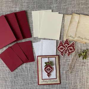 Pode incluir: Um conjunto de materiais para artesanato para fazer cart&otilde;es de Natal. O conjunto inclui cartolina vermelha, cartolina branca, cartolina creme, cartolina marrom, um cart&atilde;o branco com um design de enfeite vermelho e tr&ecirc;s enfeites vermelhos com designs brancos. Os enfeites t&ecirc;m o texto "Merry Christmas".