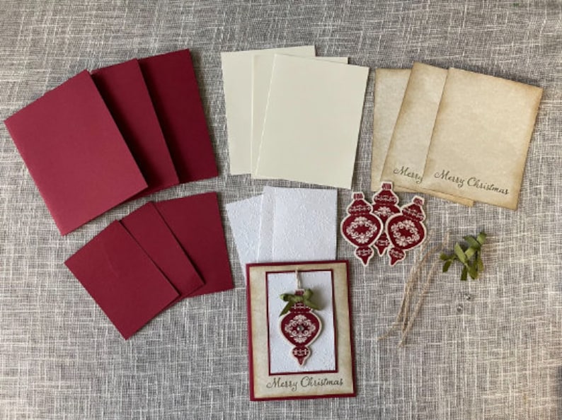 Pode incluir: Um conjunto de materiais para artesanato para fazer cart&otilde;es de Natal. O conjunto inclui cartolina vermelha, cartolina branca, cartolina creme, cartolina marrom, um cart&atilde;o branco com um design de enfeite vermelho e tr&ecirc;s enfeites vermelhos com designs brancos. Os enfeites t&ecirc;m o texto "Merry Christmas".