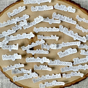 May include: A collection of white stickers with various messages written in black text. The messages include "Feel Better", "Get Well Soon", "Just For You", "You're In My Thoughts", "Baby Boy", "Make a Wish", "Aged To Perfection", "To The Newlyweds", "Happy Grad", "Thinking of", "Welcome, Baby", "Baby Girl", "Happy Anniversary", "Surprise", "Thanks So Much", "Congratulations", "Missing You", "Birthday Wishes", "Celebrating You", "Just Because", "I Love", "Hello, Friend", "Best Wishes", "From All of Us", "You're So Wonderful", "For Your Shower", and "Thank You".