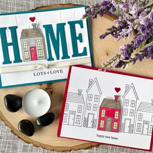 May include: Two handmade greeting cards with a house theme. The first card is teal blue with the word "HOME" in white letters with a small house in the middle. The second card is red with a row of five houses, one of which is red and the rest are white. The text "lots of love" is on the first card and "happy new home" is on the second card.