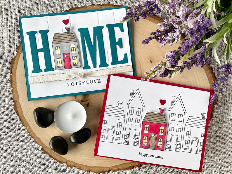 May include: Two handmade greeting cards with a house theme. The first card is teal blue with the word "HOME" in white letters with a small house in the middle. The second card is red with a row of five houses, one of which is red and the rest are white. The text "lots of love" is on the first card and "happy new home" is on the second card.