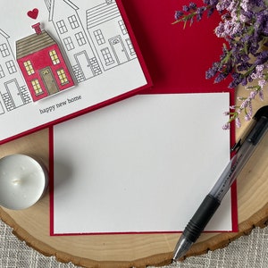 May include: A white card with a red border and a drawing of houses with a red house in the center. The card says "happy new home". The card is on a wooden surface with a white candle and a pen.