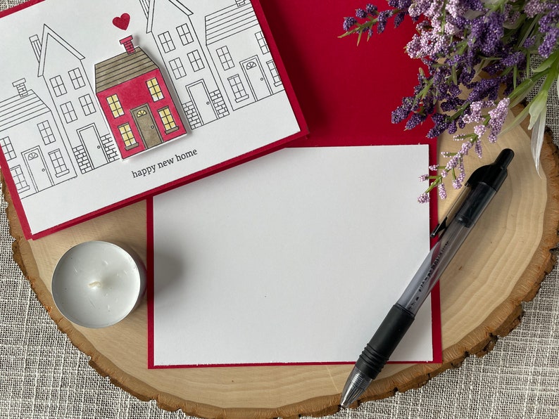 May include: A white card with a red border and a drawing of houses with a red house in the center. The card says "happy new home". The card is on a wooden surface with a white candle and a pen.