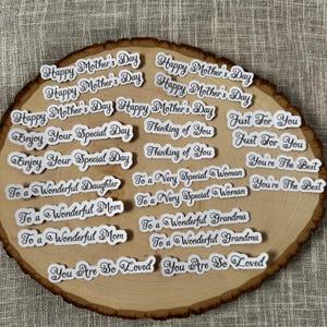 May include: A collection of white die-cut stickers with black script on a wooden slice. The stickers feature phrases like "Happy Mother's Day," "Enjoy Your Special Day," and "You're The Best." The wooden slice has a natural, light brown color.
