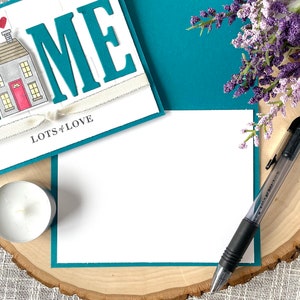May include: A teal card with the word "HOME" in large white letters, a small house illustration, and the text "Lots of Love". The card is on a wooden surface with a white candle and a black pen.