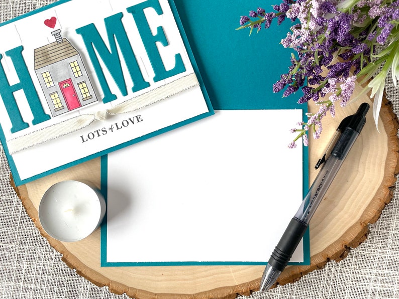 May include: A teal card with the word "HOME" in large white letters, a small house illustration, and the text "Lots of Love". The card is on a wooden surface with a white candle and a black pen.