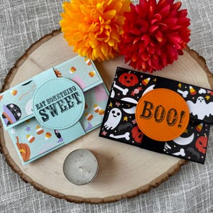 May include: Two Halloween-themed gift card holders on a wooden surface. One is light blue with candy corn and pumpkin patterns, the other is black with ghosts and bones. Both have circular accents with text. Orange and red pom-pom flowers are in the background.