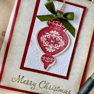 Pode incluir: Um cart&atilde;o de Natal branco com borda vermelha, decorado com um enfeite de Natal vermelho e branco com um la&ccedil;o verde e o texto "Merry Christmas".