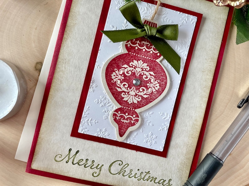 Pode incluir: Um cart&atilde;o de Natal branco com borda vermelha, decorado com um enfeite de Natal vermelho e branco com um la&ccedil;o verde e o texto "Merry Christmas".
