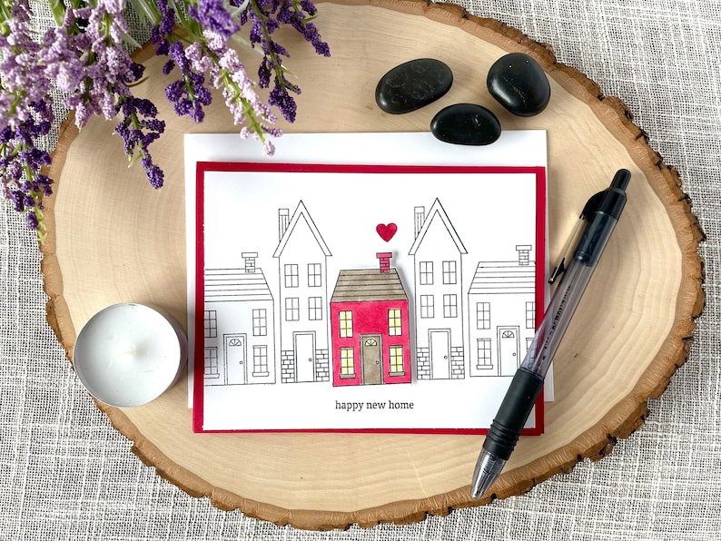 May include: A red and white card with a drawing of a row of houses. The middle house is red and has a heart above it. The text "happy new home" is below the houses.