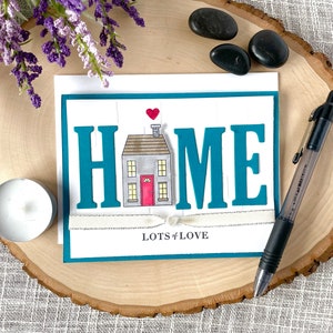 May include: A teal card with the word "HOME" in large, white letters. A small house illustration is in the center of the word. The house is gray with a red door and a white heart above it. The card has a white ribbon tied around it and the words "Lots of Love" are printed below the house.