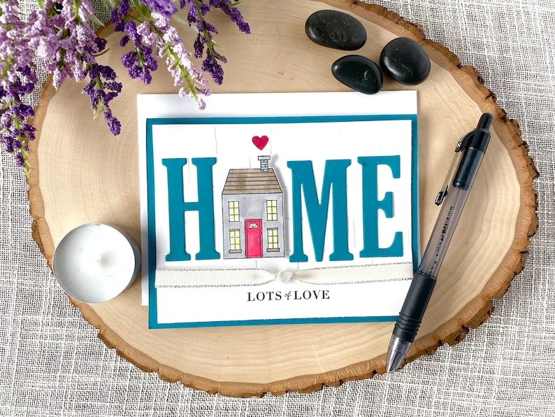 May include: A teal card with the word "HOME" in large, white letters. A small house illustration is in the center of the word. The house is gray with a red door and a white heart above it. The card has a white ribbon tied around it and the words "Lots of Love" are printed below the house.