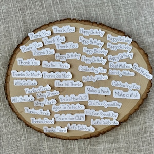 May include: A collection of white stickers with various messages, including "Thank You", "Happy Birthday", "Best Wishes", "Celebrating You", "Make a Wish", "It's Your Special Day", "You're How Old?!", "Aged To Perfection", "Let's Celebrate", and "Thanks, Friend". The stickers are arranged on a wooden surface.