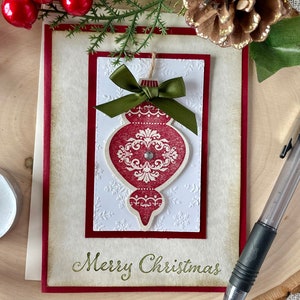 Pode incluir: Um cart&atilde;o de Natal com um ornamento vermelho e branco com um la&ccedil;o verde. O cart&atilde;o tem um fundo branco com uma borda vermelha e as palavras "Feliz Natal" escritas em verde.
