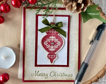 May include: A Christmas card featuring a red and white bauble with a green bow. The card has a white background with a red border and the words "Merry Christmas" written in green.