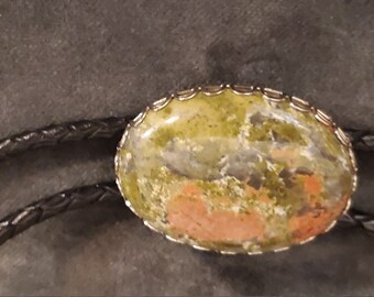 May include: A bolo tie with a green, brown, and orange stone set in a silver-toned oval frame. The bolo tie has a black leather cord.