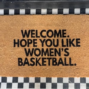 May include: A rectangular tan coir doormat with a black and white checkered border. The mat features the black text: WELCOME. HOPE YOU LIKE WOMEN'S BASKETBALL. The text is centered in four lines, with a period after each word or phrase.