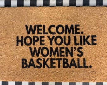 May include: A rectangular tan coir doormat with a black and white checkered border. The mat features the black text: WELCOME. HOPE YOU LIKE WOMEN'S BASKETBALL. The text is centered in four lines, with a period after each word or phrase.