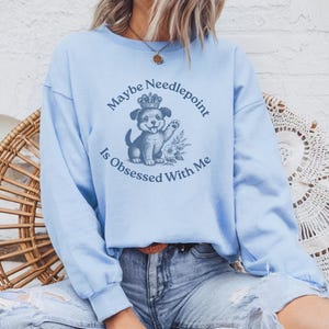 May include: Light blue sweatshirt with the text "Maybe Needlepoint is Obsessed With Me" and a graphic of a puppy wearing a crown. The sweatshirt has long sleeves and a crew neck. The puppy graphic is in the center of the sweatshirt.