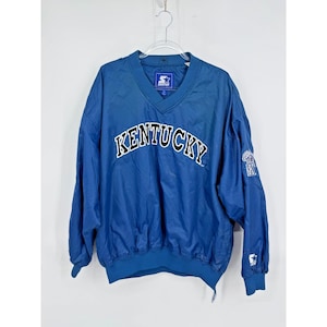 May include: A blue Starter pullover jacket with a V-neck collar. The word "KENTUCKY" is arched across the chest in white and black lettering. A logo is on the left sleeve and the Starter logo is on the collar.