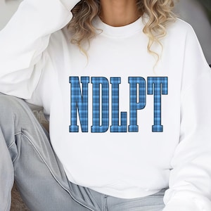 May include: White sweatshirt with the letters "NDLPT" in a blue plaid pattern. The letters are in a bold, block font. The sweatshirt has long sleeves and a crew neck.