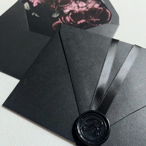 May include: Black envelopes with a floral interior design. One envelope is open, revealing a pink and black floral pattern. The other envelope is sealed with a black wax seal and black ribbon. The wax seal has an embossed letter "R".