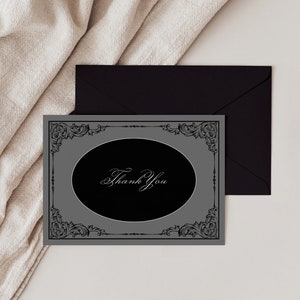 May include: A gray thank you card with a black oval in the center. The oval has a white cursive inscription that says "Thank You". The card is framed with a black ornate design.