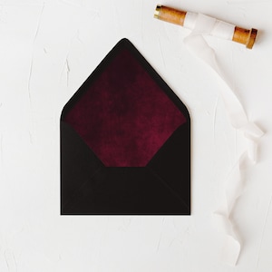 May include: A black envelope with a burgundy velvet inner lining. The envelope is lying flat on a white textured surface. A spool of white ribbon is lying on the surface to the right of the envelope.