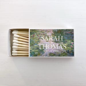 May include: A rectangular matchbox with a sliding tray containing wooden matches with white tips. The cover features a water lily painting print with the white text SARAH & THOMAS and the date 07.24.2028 printed in the center of the design.