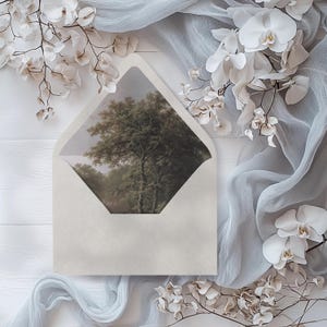May include: A white envelope with a window showing a watercolor painting of a forest scene. The envelope is surrounded by white flowers and a light blue fabric.