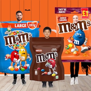 May include: Three people dressed in M&M's candy costumes for Halloween. The costumes are designed to look like large bags of M&M's. The bags are blue, brown, and orange, with the M&M's logo and various flavors displayed. The background is orange with Halloween decorations.