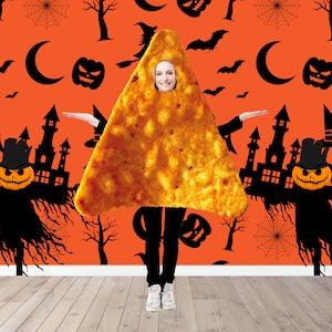 May include: A woman dressed as a nacho chip for Halloween. She is standing in front of a wall with a Halloween theme. The wall is orange with black silhouettes of bats, spiders, and pumpkins.