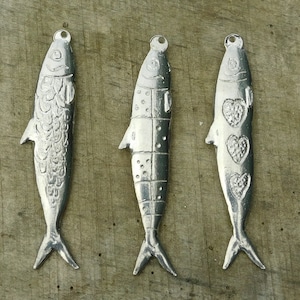 Pode incluir: Três pingentes de peixe prateados com diferentes designs. Os pingentes são detalhados e têm um acabamento brilhante.