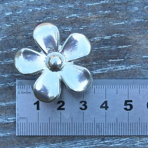 May include: A silver-coloured metal flower charm with five petals and a centre bead. The charm is approximately 3 centimetres wide.