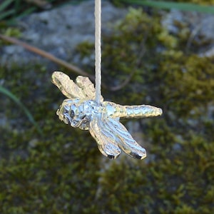 May include: A silver dragonfly charm hanging from a white string. The dragonfly is detailed with intricate wings and a textured body.