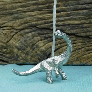 Puede incluir: Una figurita de dinosaurio de metal plateado con cuello y cola largos. El dinosaurio está de pie sobre una superficie azul.