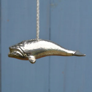 Op de afbeelding: Een zilveren walvisvormige lichtschakelaar. De walvis hangt aan een wit koord.