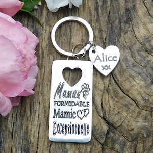 Może przedstawiać: Srebrny brelok do kluczy z prostokątnym zawieszką, na której widnieje napis "Maman formidable Mamie exceptionnelle" z wycięciem w kształcie serca i małym wzorem kwiatu. Do breloka dołączona jest mniejsza zawieszka w kształcie serca z imieniem "Alice".