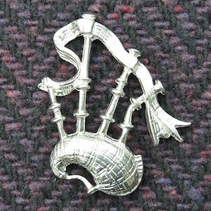 Könnte beinhalten: Eine silberfarbene Dudelsack-Anstecknadel mit einem karierten Muster. Die Nadel ist ein traditionelles schottisches Symbol.