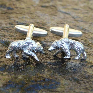 May include: A pair of silver cufflinks featuring a detailed bear design. Each cufflink has a bear walking on all fours, with a textured surface.