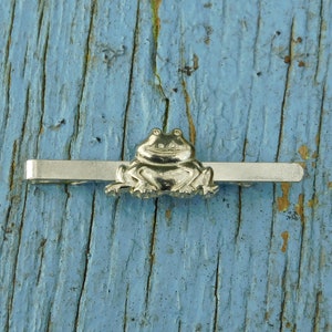 May include: A silver tie bar featuring a frog design. The frog is sitting on a silver bar.
