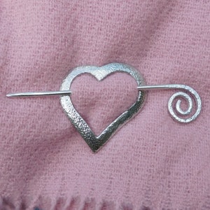 Puede incluir: Broche de chal de plata en forma de corazón con un detalle en espiral. La broche está sobre un fondo de tela rosa.